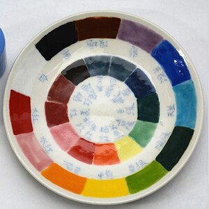 K&ouml;nnte beinhalten: Ein wei&szlig;er Keramikteller mit einem farbenfrohen Farbkreis-Design. Das Rad zeigt ein Spektrum an Farbt&ouml;nen, darunter Rot, Gelb, Blau und Gr&uuml;n, mit chinesischen Schriftzeichen. Der Teller ist rund und hat eine gl&auml;nzende Oberfl&auml;che.