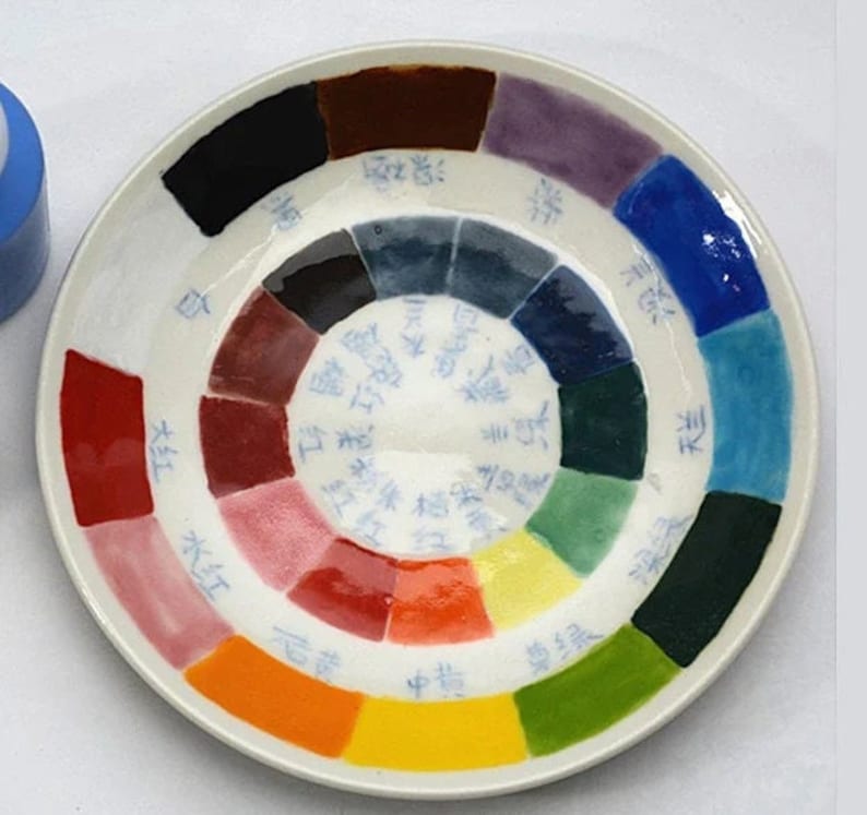 K&ouml;nnte beinhalten: Ein wei&szlig;er Keramikteller mit einem farbenfrohen Farbkreis-Design. Das Rad zeigt ein Spektrum an Farbt&ouml;nen, darunter Rot, Gelb, Blau und Gr&uuml;n, mit chinesischen Schriftzeichen. Der Teller ist rund und hat eine gl&auml;nzende Oberfl&auml;che.