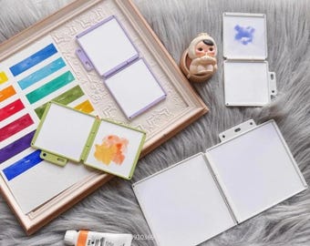 May include: A collection of watercolor palettes in various colors, including purple, green, and white. The palettes are open and closed, with some containing watercolor paint. A tube of orange paint is also visible.