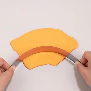 May include: A curved metal blade tool is pressed into a flat, irregular piece of bright orange modelling clay. The thin, silver-coloured cutter has a smooth, arc-shaped edge designed for shaping or slicing through the pliable, soft material.