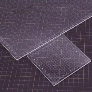 May include: Two clear acrylic cutting mats with grid lines and measurement markings. One mat is rectangular, the other is square. The mats have measurements in inches and centimeters. The background is a dark purple cutting mat.