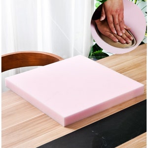 May include: A pink square foam cushion, approximately 38 cm by 38 cm, sits on a wooden surface. The cushion is thick and appears to be soft and comfortable.