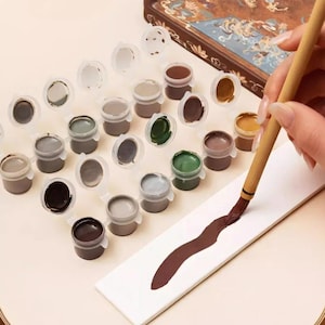 Puede incluir: Un conjunto de pequeños botes de pintura circulares de plástico con tapas abiertas, que contienen varios tonos de pintura acrílica, incluyendo marrón, verde, gris y blanco. Un pincel de madera aplica un trazo de pintura marrón oscuro.