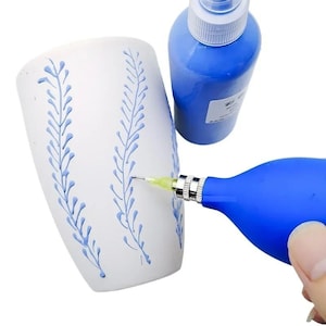 Pode incluir: Uma caneca de cerâmica branca decorada com padrões de folhas azuis verticais, posicionada ao lado de um frasco de tinta azul e uma ferramenta aplicadora de precisão azul com uma ponta metálica fina para aplicar a tinta na caneca.