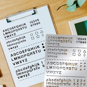 Könnte beinhalten: Ein weißes Klemmbrett und eine silberne Schablone mit schwarzen Groß- und Kleinbuchstaben, Zahlen und Symbolen. Auf dem Klemmbrett sind das Alphabet und Zahlen abgebildet. Die Schablone dient zum Erstellen von Buchstaben und Zahlen.