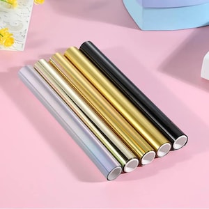 Puede incluir: Cinco rollos de papel de regalo metálico en plata, dorado y negro. Los rollos están dispuestos en diagonal sobre una superficie rosa, con los extremos de los rollos visibles, mostrando un interior blanco. El papel tiene una superficie brillante y reflectante.