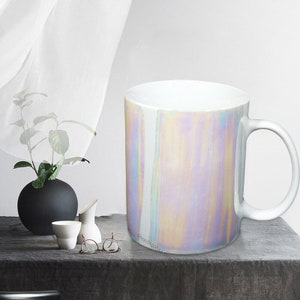 May include: A white ceramic mug with a iridescent finish. The mug has a handle and is sitting on a gray surface.