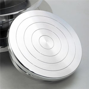 Puede incluir: Una plataforma giratoria para pasteles de plata con círculos concéntricos en la superficie. La plataforma giratoria está hecha de metal y tiene un acabado liso y pulido.