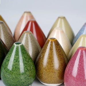 Puede incluir: Una colección de pequeños jarrones de cerámica en forma de lágrima de varios colores, incluyendo verde, marrón, rojo, beige y azul. Los jarrones tienen un esmalte moteado y un acabado brillante.
