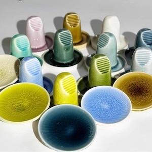 May include: A collection of colorful ceramic thimbles and matching saucers in various shades of blue, green, yellow, and pink. The thimbles have a ribbed design on the top. The saucers have a textured, crackle glaze.
