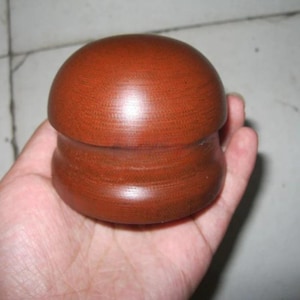 Puede incluir: Un tapón de botella de madera marrón con una parte superior redondeada y una base cilíndrica. El tapón se sostiene en una mano y parece estar hecho de madera pulida. La veta de la madera es visible en la superficie.