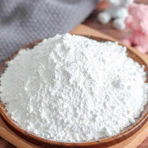 May include: A wooden bowl filled with a large quantity of white powder. The powder has a fine, textured appearance. The bowl is set on a wooden surface, with a grey cloth visible in the background.