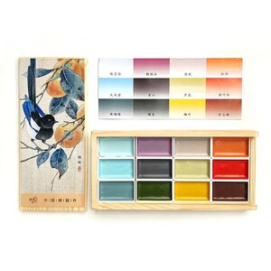 May include: A watercolor paint set with a wooden box containing 12 colorful paint pans. A color chart displays various hues. The set includes a decorative box with a bird and fruit illustration.
