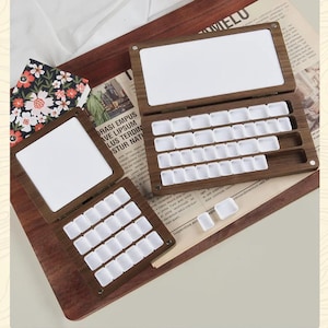 Może przedstawiać: Drewniane palety akwarelowe z białymi studzienkami na farby i białą powierzchnią do mieszania. Palety są otwarte i zamknięte, prezentując design. Drewno ma naturalne usłojenie, a palety ułożone są na drewnianej tacy.