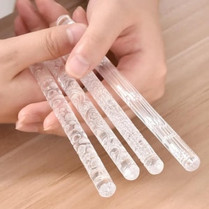 May include: Four clear, textured rolling pins with decorative patterns. The rolling pins are held in a person's hands. The rolling pins have different patterns, including floral and linear designs. The rolling pins are made of a clear material.