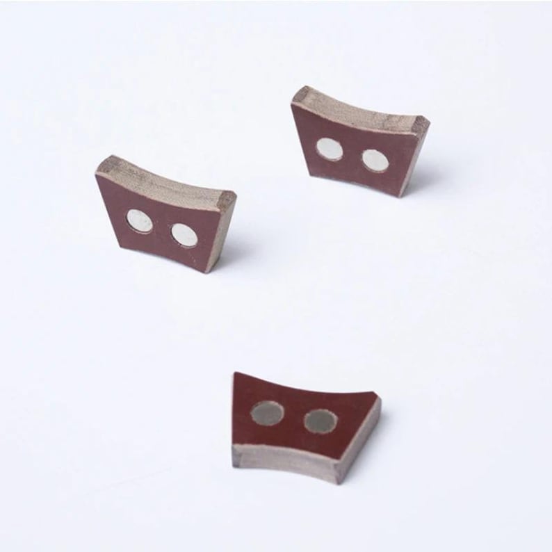 Pu&ograve; includere: Tre piccoli oggetti curvi a forma di trapezio. Ognuno ha una faccia rosso scuro con due inserti circolari color argento. I bordi sono di un colore pi&ugrave; chiaro e neutro. Gli oggetti sono disposti su uno sfondo bianco.