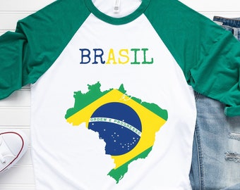 May include: A white t-shirt with green and blue raglan sleeves. The shirt features a graphic of the Brazilian flag in the shape of the country's outline. The text "BRASIL" is printed above the flag.