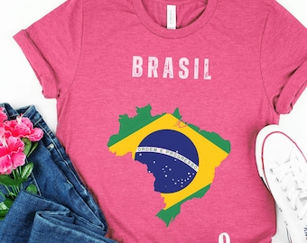 May include: A pink t-shirt with a graphic of the outline of Brazil with the Brazilian flag inside. The text "BRASIL" is printed in white above the graphic. The shirt is laid out on a white surface with a pair of blue jeans and white sneakers.