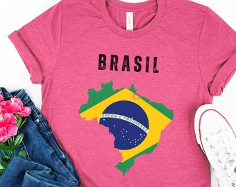 May include: A pink t-shirt with the word "BRASIL" printed in black on the front. The shirt also features a graphic of the Brazilian flag in the shape of the country's outline. The flag is green, yellow, and blue with the words "ORDEM E PROGRESSO" written on a blue band.