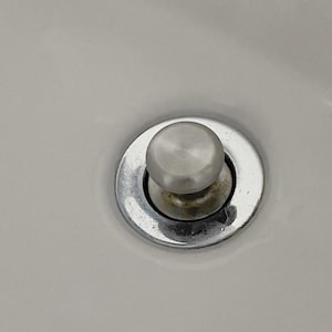 May include: A close-up of a chrome sink drain stopper with a round, brushed metal knob. The drain stopper is set in a white sink basin. The drain stopper has some discoloration.