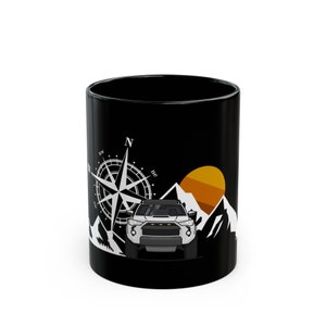 May include: Black ceramic mug with a white compass rose, a white SUV, and a mountain range silhouette with an orange sun. The mug is a standard 325 ml size.