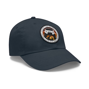 Puede incluir: Gorra de béisbol azul marino con un parche circular que muestra un vehículo todoterreno blanco y el texto "Yours Can Go Faster, Mine Can Go Anywhere". El parche tiene detalles marrones, naranjas y blancos. La gorra es de tela suave.