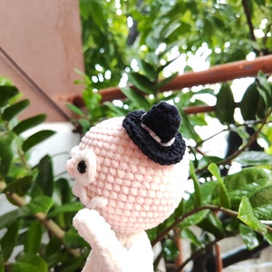 May include: A pink and white crocheted amigurumi doll wearing a black hat with a white band. The doll has large, black eyes and a white smile. The doll is standing on a pink surface.