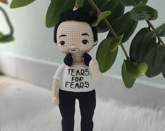 May include: A crocheted doll with black hair, wearing a white shirt with the text "Tears for Fears" and black pants. The doll is standing on a white fluffy surface.