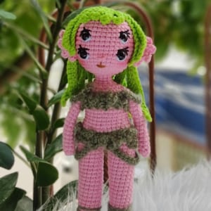 May include: A pink crocheted doll with green hair and four eyes. The doll is wearing a green and brown outfit and is standing on a wooden base.