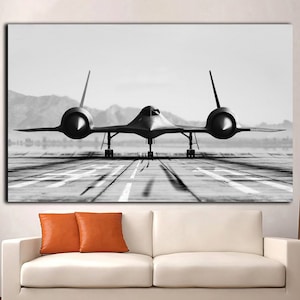 Op de afbeelding: Een zwart-witfoto van een gestroomlijnd, zwart straalvliegtuig op een landingsbaan. Het vliegtuig is gericht op de camera en heeft het landingsgestel naar beneden. De achtergrond is een wazig beeld van bergen.