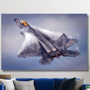 Op de afbeelding: Een grijs en zwart gevechtsvliegtuig vliegt door de wolken en laat een rookstreep achter.