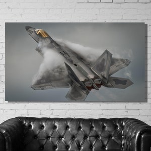 Op de afbeelding: Een canvas print van een grijze F-22 Raptor straaljager in vlucht, van onderaf gezien. De straaljager staat omhoog gekanteld, met dampsporen die uit de vleugels komen. De cockpit is goudkleurig geaccentueerd.