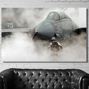 Op de afbeelding: Een militair straalvliegtuig dat taxiet op een landingsbaan, met een piloot in een helm en vluchtpak dat achter hem loopt. Het vliegtuig is omgeven door een wolk van stoom of rook.