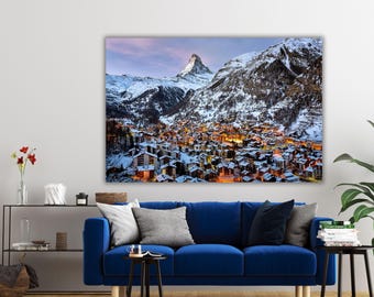May include: A rectangular wall art print featuring a snowy mountain landscape at dusk, showcasing the Matterhorn peak towering over a village with glowing yellow lights, set against a soft pink and blue sky above a deep blue sofa.
