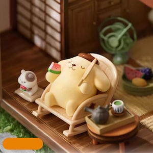 K&ouml;nnte beinhalten: Eine Miniatur-Diorama-Szene mit einer gelben Cartoon-Hundefigur, die sich in einem Schaukelstuhl entspannt und eine Wassermelone h&auml;lt. Eine kleine Hundefigur sitzt in der N&auml;he. Ein Miniatur-Teeservice und ein Ventilator sind ebenfalls vorhanden.