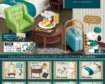 May include: A miniature Snoopy's Hotel Life diorama set. The set includes a green armchair, a round table with a meal, a bed, and a bellhop Snoopy figure. The set also includes a blue suitcase on a luggage cart. The packaging features the Peanuts logo.