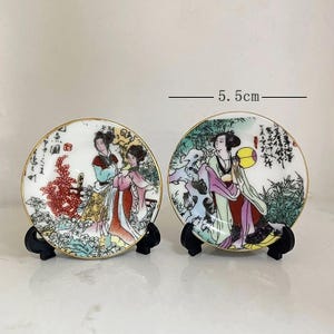 Op de afbeelding: Twee kleine, ronde decoratieve borden, elk 5,5 cm in diameter, met gedetailleerde illustraties van vrouwen in traditionele Aziatische kleding. De borden hebben gouden randen en worden op zwarte standaards gepresenteerd. Het kunstwerk bevat levendige kleuren en ingewikkelde details.