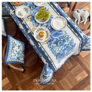 Op de afbeelding: Een blauwe en witte bloemen tafelkleed met een blauwe rand. Het tafelkleed ligt op een houten tafel met een wit bord, een blauw en wit bord en een kom met druiven. Er zijn ook twee stoelen met blauwe en witte bloemen kussens.
