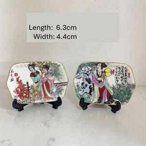 Może przedstawiać: Dwa małe dekoracyjne talerzyki z pozłacanym brzegiem, każdy z ręcznie malowanym obrazkiem przedstawiającym kobiety w tradycyjnych japońskich strojach. Talerzyki mają 6,3 cm długości i 4,4 cm szerokości.