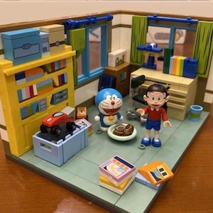 May include: A detailed miniature diorama of Doraemon and Nobita's room. The scene includes a yellow bookcase, a desk, and various accessories. The figures are positioned in a room setting with blue and green accents.