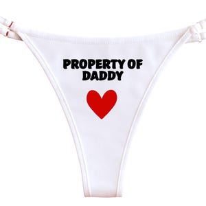 Puede incluir: Tanga blanco con la frase "PROPERTY OF DADDY" en negro y un corazón rojo. El tanga tiene tirantes finos y un panel frontal triangular.