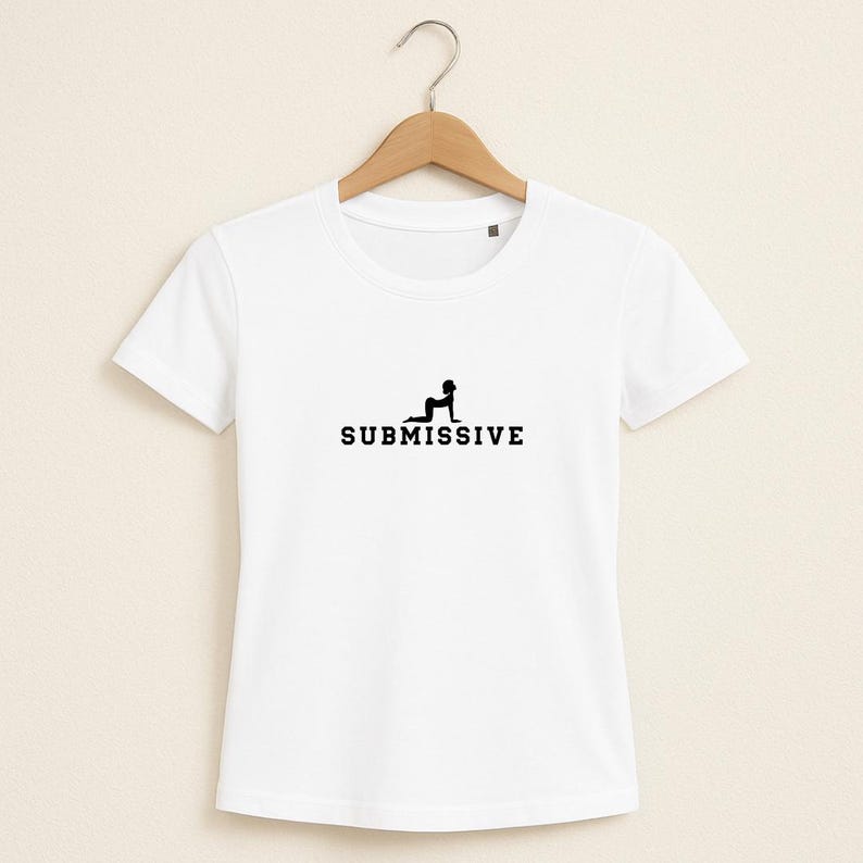 Może przedstawiać: Biała koszulka z napisem "SUBMISSIVE" w kolorze czarnym, z sylwetką osoby klęczącej nad tekstem. Koszulka wisi na drewnianym wieszaku na jasnobeżowym tle.