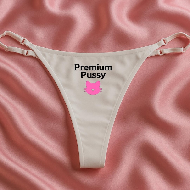 May include: Cream-coloured thong underwear with delicate straps. The words "Premium Pussy" are printed in black, with a pink cat face graphic. The underwear is displayed on a pink satin surface.