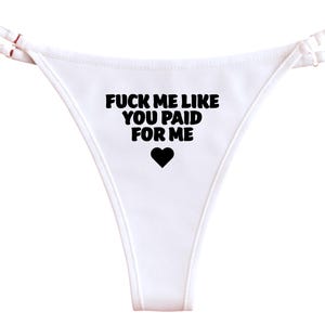 May include: White thong underwear with black text that reads "FUCK ME LIKE YOU PAID FOR ME" and a black heart symbol. The straps are thin and white.