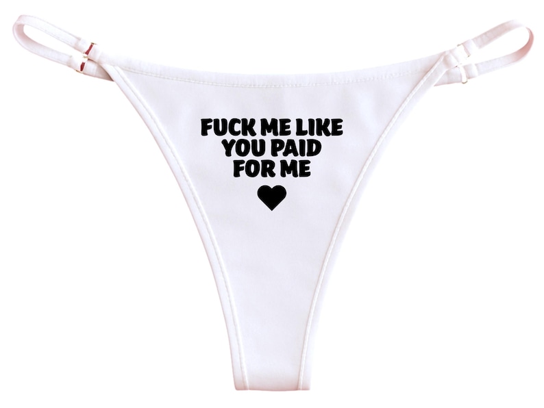 May include: White thong underwear with black text that reads "FUCK ME LIKE YOU PAID FOR ME" and a black heart symbol. The straps are thin and white.
