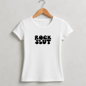 May include: A white t-shirt with a crew neck and short sleeves. The shirt features the words "ROCK SLUT" in bold, black, bubble letters across the chest. The shirt is hanging on a wooden hanger.