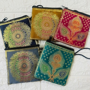 Puede incluir: Una colección de pequeñas bolsas de tela cuadradas con intrincados diseños bordados en oro y color. Cada bolsa tiene una cremallera negra y una correa de muñeca a juego. Las bolsas presentan patrones de mandala y plumas de pavo real.