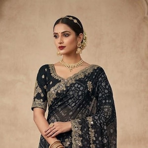 May include: A woman wearing a black saree with gold embroidery and a matching blouse. The saree features a delicate pattern and a sheer border. The woman is also wearing gold jewelry, including a necklace, earrings, and bangles. The background is a neutral color.