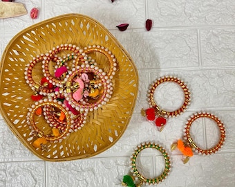 May include: A collection of colorful bangles with pearl accents and tassel charms. The bangles are arranged in a woven basket on a white brick background.
