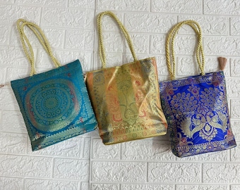 May include: Three decorative tote bags with gold rope handles. The bags feature intricate patterns in teal, gold, and blue. The teal bag has a circular design, the gold bag has a floral pattern, and the blue bag has a peacock design.